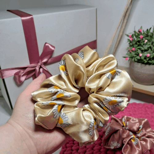 Gumka do włosów w kolorze beżowym i motywem stokrotek trzymana na ręce, w tle ozdobne pudełko i kwiat