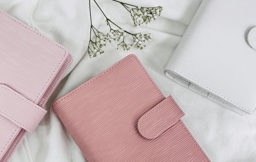 Trzy eleganckie organizery w pastelowych odcieniach różu i bieli, wykonane z tłoczonej ekoskóry, leżące na białym tle z dekoracyjnymi gałązkami gipsówki.